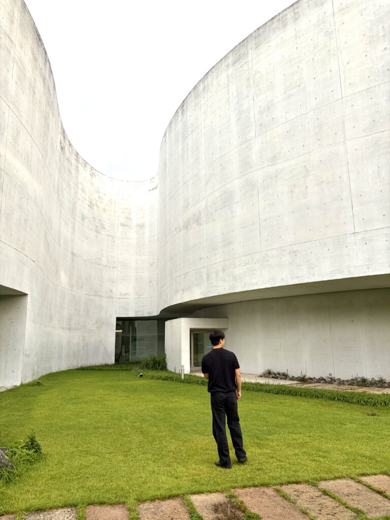 Visitor standing on a green lawn beneath the tall curved concrete walls of Mimesis Art Museum in Paju